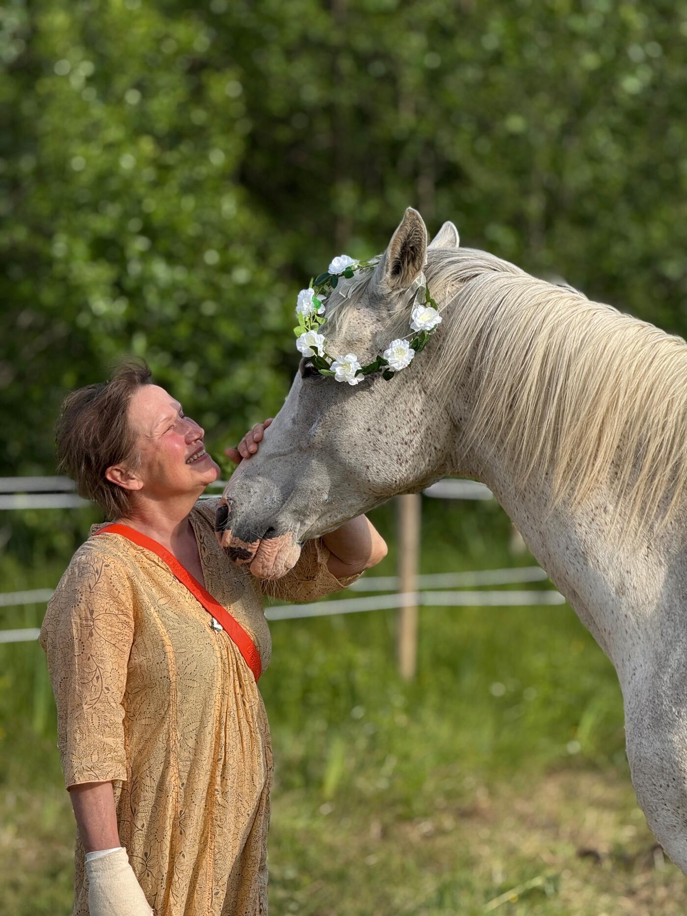 Annika håller om mulen på sin vita häst, som har en blomsterkrans på huvudet, framför sommargröna träd