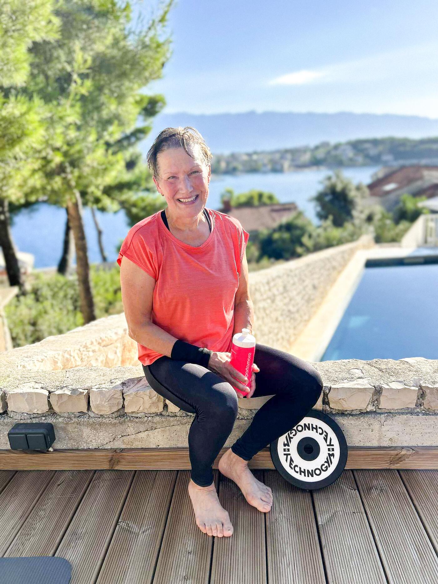 Annika sitter leende på en stenmur med en vattenflaska i handen, efter träning med havet i bakgrunden